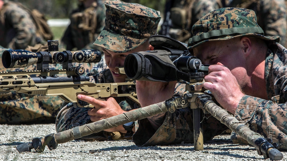 Two Marines lying behind weapons squint while looking through weapon scopes.