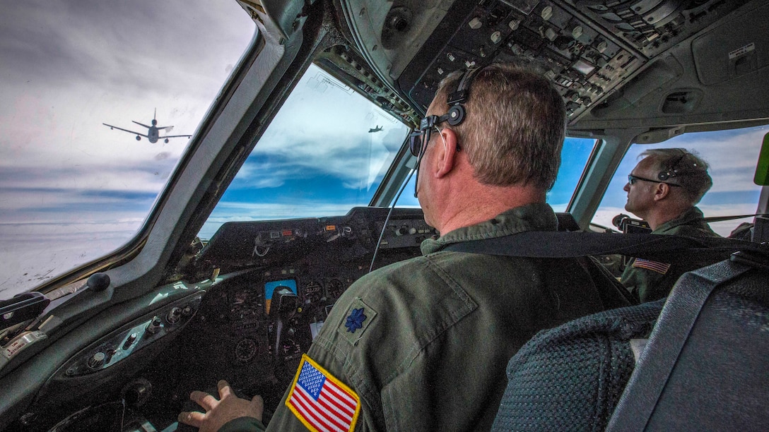 Two pilots look out of a cockpit window at an aircraft flying nearby.