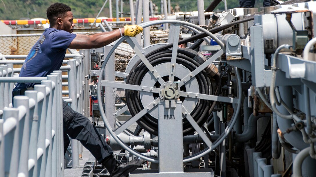A sailor, shown in profile, pulls a large grey hose reel.