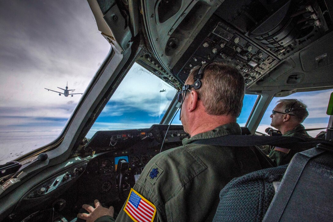 Two pilots look out of a cockpit window at an aircraft flying nearby.