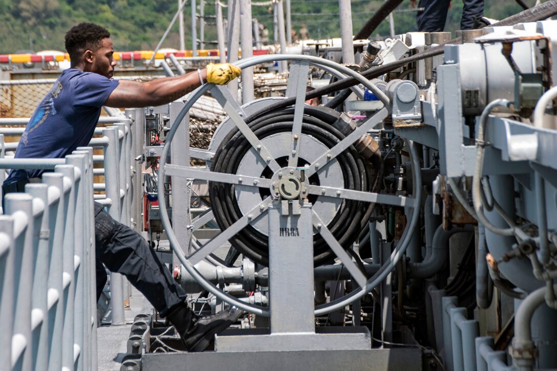 A sailor, shown in profile, pulls a large grey hose reel.