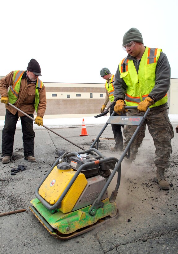 Airmen and civilians use a plate compactor to repair potholes.