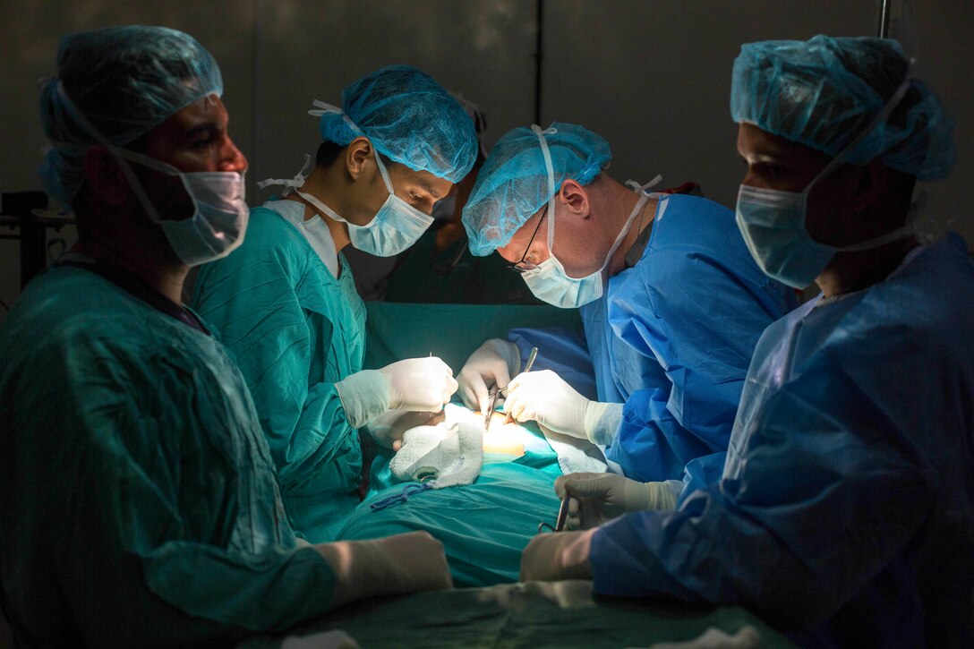 Two doctors in scrubs operate on either side of a patient, illuminated by soft light, as two others stand by.