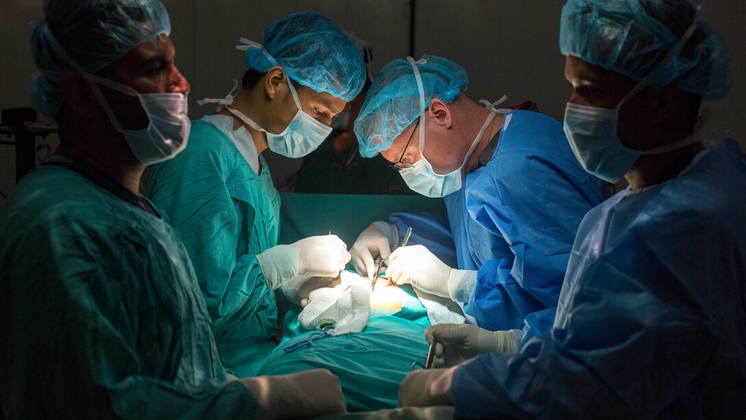 Two doctors in scrubs operate on either side of a patient, illuminated by soft light, as two others stand by.