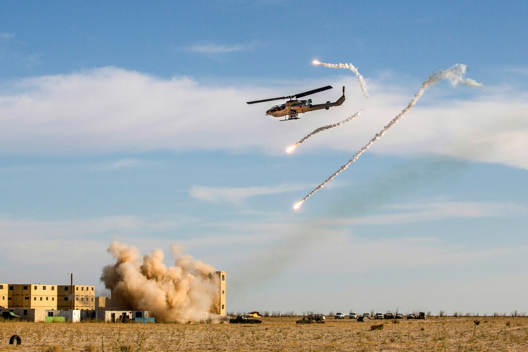 Flares shoot near a helicopter flying over a building complex partially engulfed by an explosion.