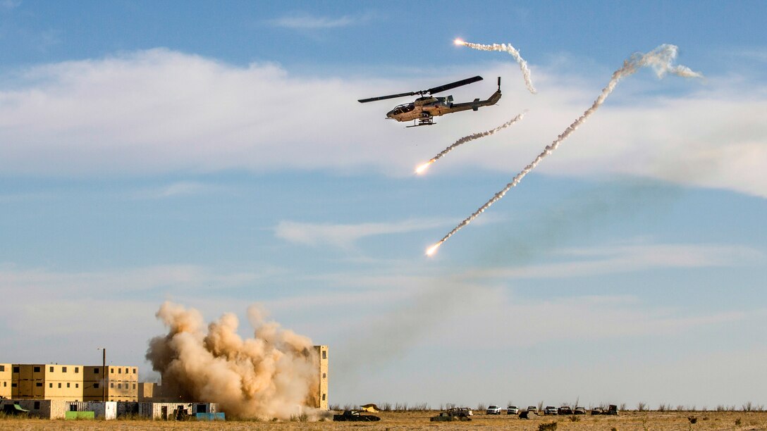 Flares shoot near a helicopter flying over a building complex partially engulfed by an explosion.
