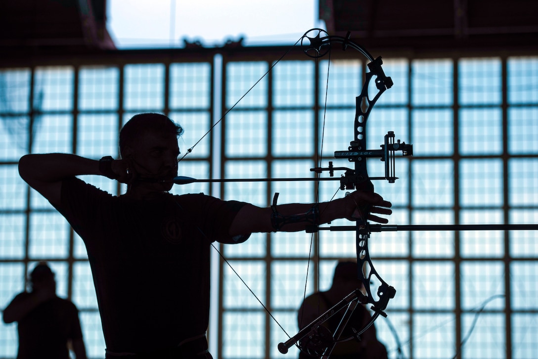 An archer draw a bow against a blue sky and windows.