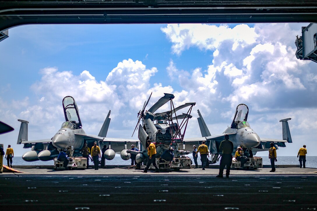 Sailors in yellow shirts walk around three fighter jets on a ship while preparing to move them.