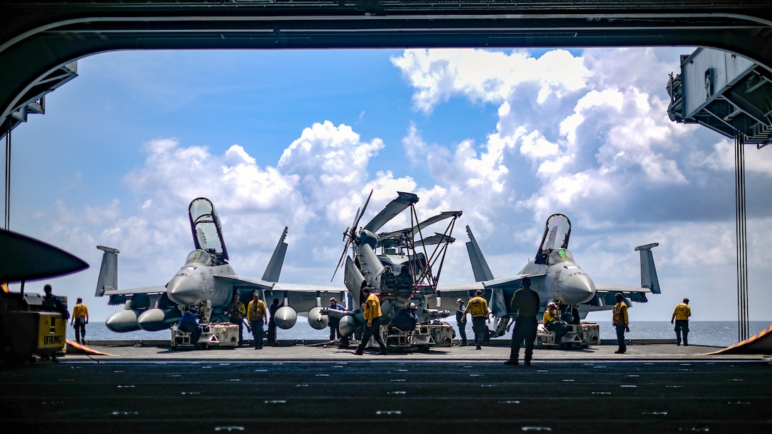 Sailors in yellow shirts walk around three fighter jets on a ship while preparing to move them.