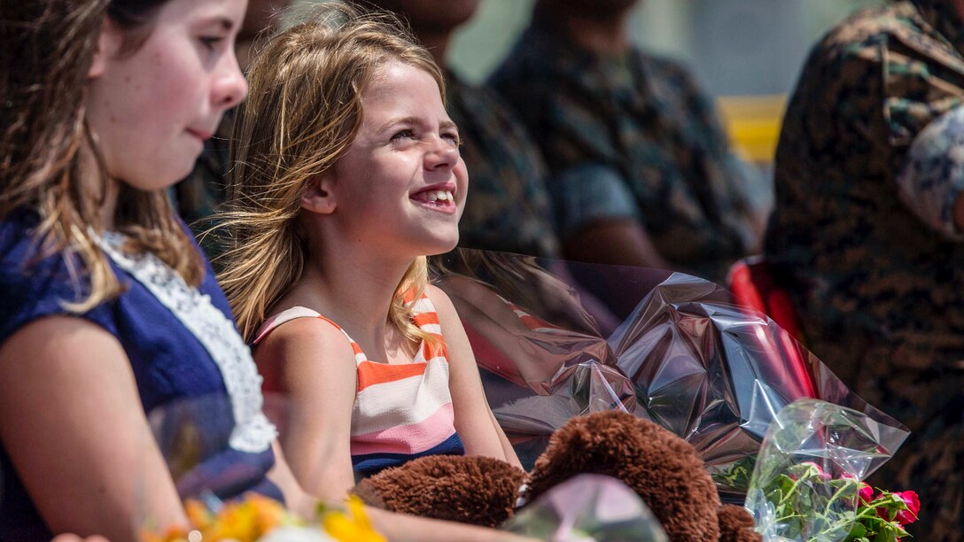 A smiling girl holds a stuffed animal and a bouquet as she sits in an audience of Marines.