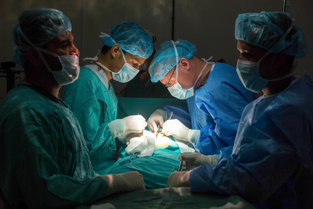 Two doctors in scrubs operate on either side of a patient, illuminated by soft light, as two others stand by.