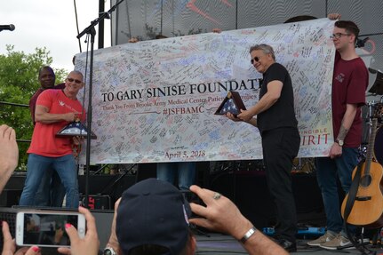 Gary Sinise and Robert Irvine pose for a photo April 5 with a banner filled with thank you messages and flags that were flown over Brooke Army Medical Center at Joint Base San Antonio-Fort Sam Houston. BAMC Commanding General Brig. Gen. Jeffrey Johnson presented the gifts to Sinise and Irvine for their continued support of the military and BAMC.