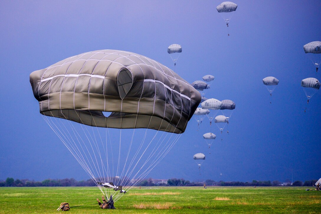 Paratroopers parachute to green grass against a blue sky.