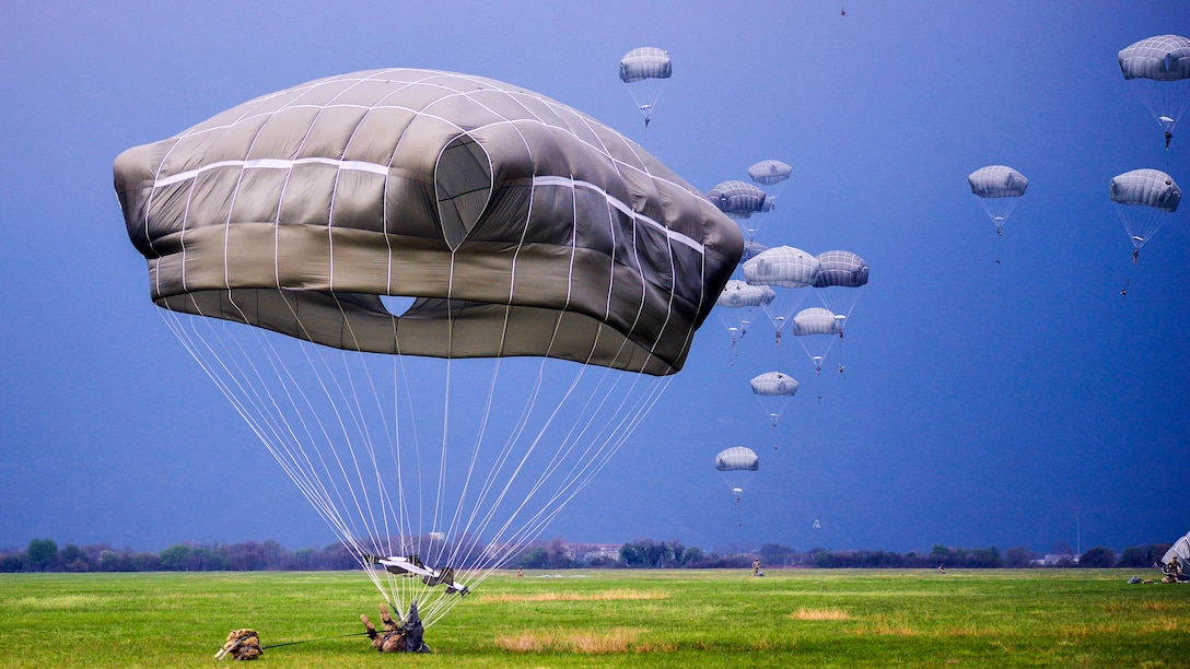 Paratroopers parachute to green grass against a blue sky.