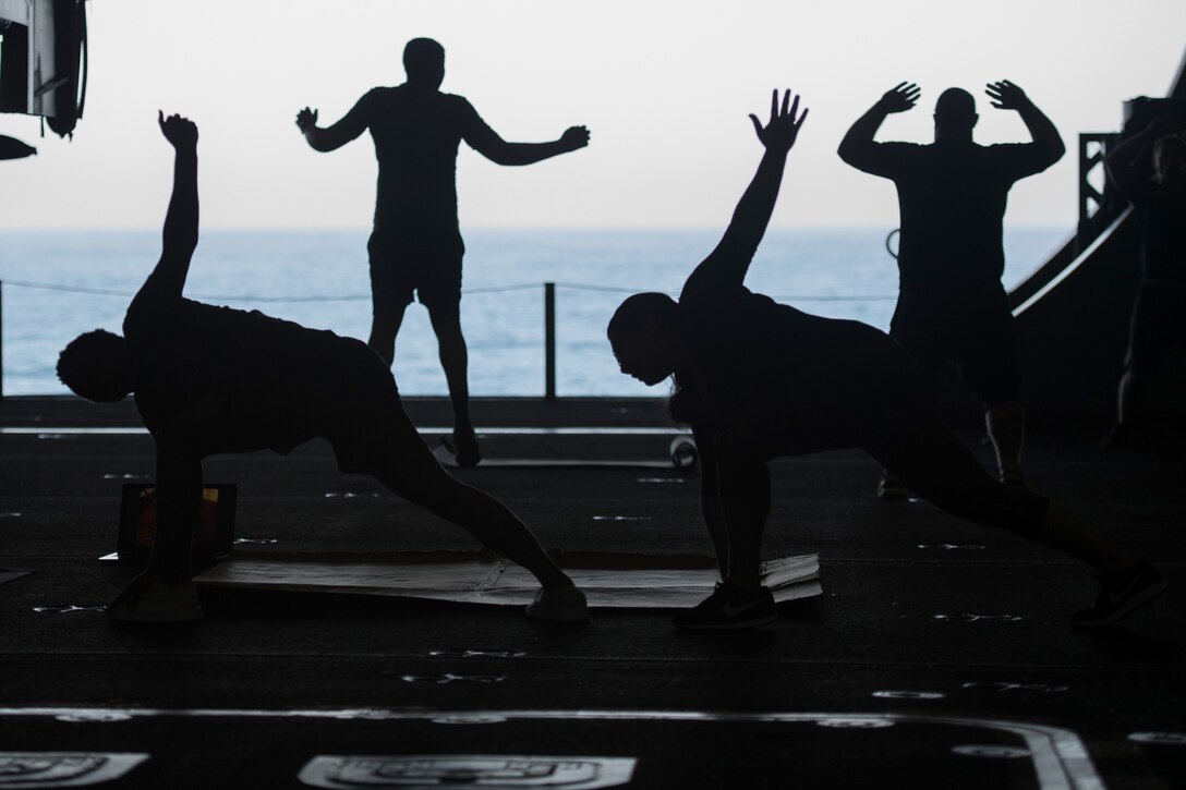 Four sailors, shown in silhouette, exercise on a ship with water in the background.
