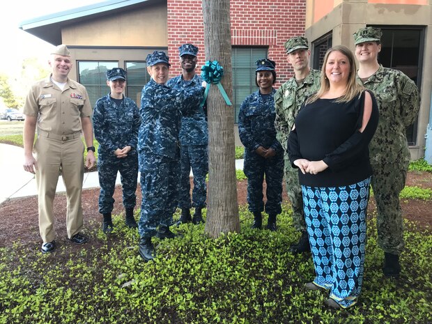 Naval Health Clinic Charleston Executive Officer Capt. Kathleen Hinz, center, along with NHCC’s Sexual Assault Prevention and Response representatives, and Ruby Godley, right, Sexual Assault Response Coordinator for Naval Weapons Station Charleston, tie a teal ribbon to a tree in front of NHCC April 2 to kick off Sexual Assault Awareness and Prevention Month. From left to right, Cdr. Robert Senko, director, NHCC Public Health; Hospitalman 3rd Class Abby Bexley, NHCC laboratory technician; Hinz; Hospitalman 2nd Class Asare Baffour, NHCC preventive medicine technician; Lt. Cdr. Crystal Bryant, NHCC family nurse practitioner; Hospitalman 2nd Class Patrick Collier, NHCC radiology technologist; Godley; Lt. Elizabeth Plowman, department head, NHCC Physical and Occupational Therapy, and NHCC’s Sexual Assault Prevention and Response Point of Contact.