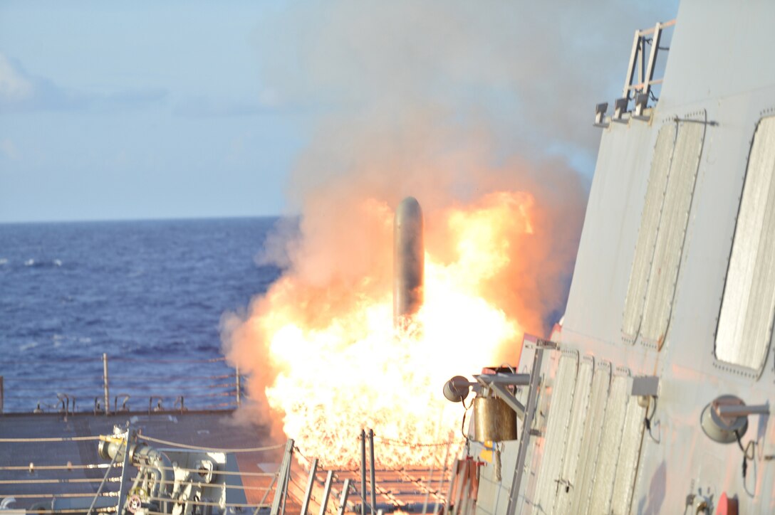 A missile emerges from a burst of flames on a ship's deck against blue sky and water.