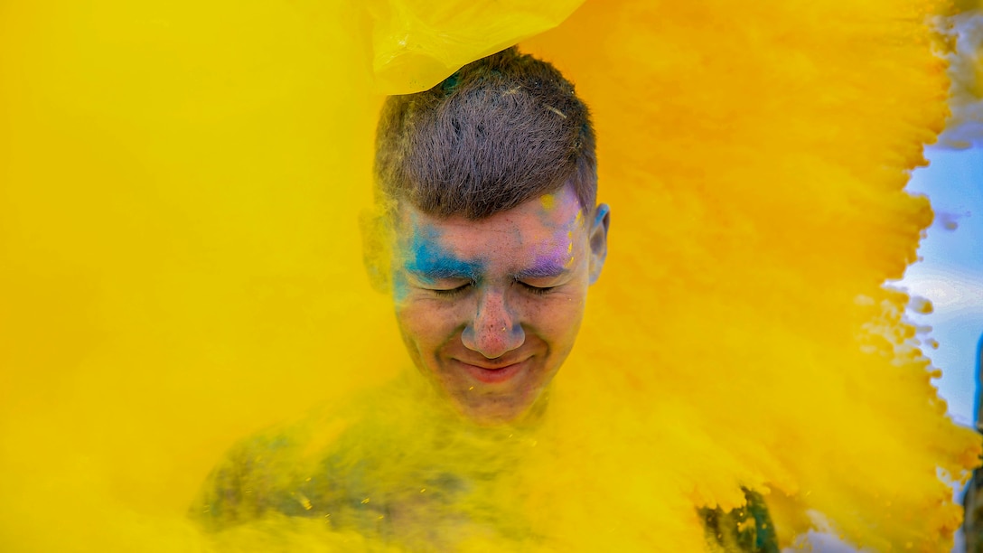 A Marine's grinning face, smeared with blue and purple powder, emerges from the center of a think cloud of yellow powder.