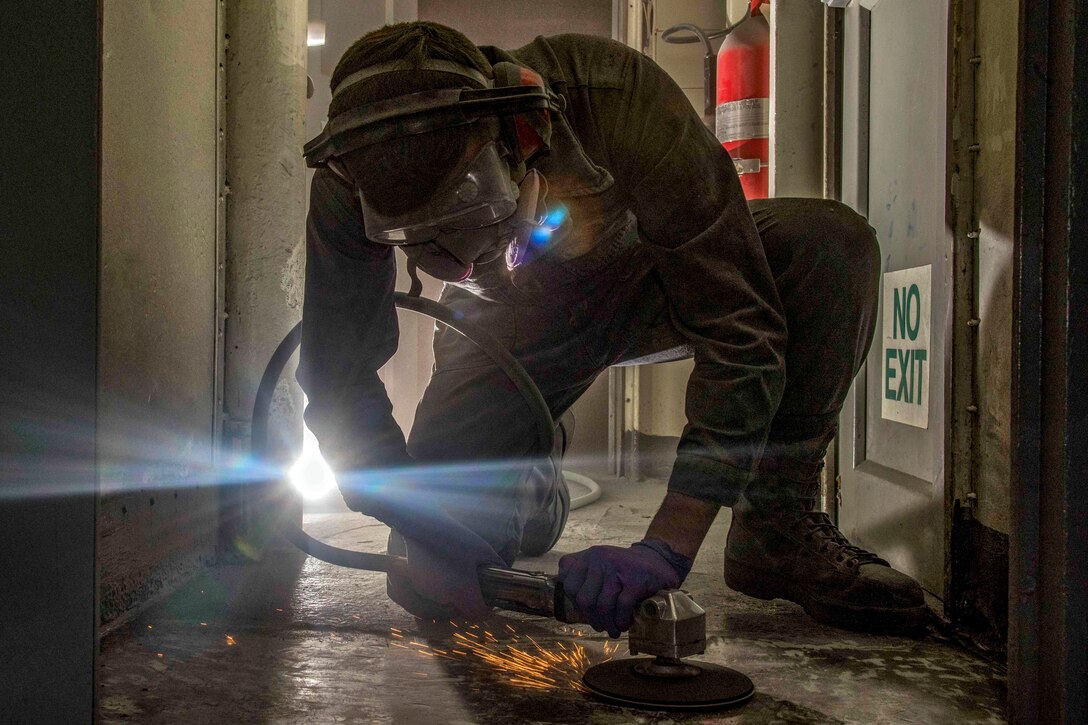 A sailor in protective head wear bends on one knee and uses a tool to grind a floor, creating sparks.