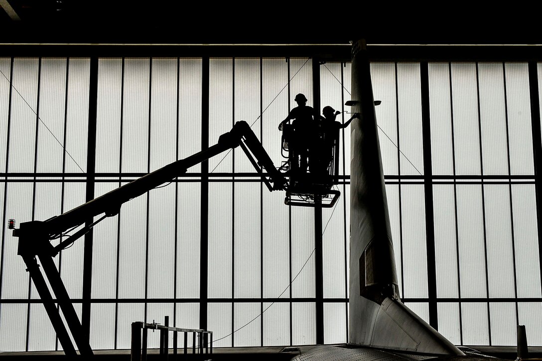Two airmen, shown in silhouette, stand on an elevated platform to conduct work on an aircraft tail.