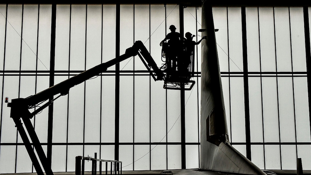 Two airmen, shown in silhouette, stand on an elevated platform to conduct work on an aircraft tail.