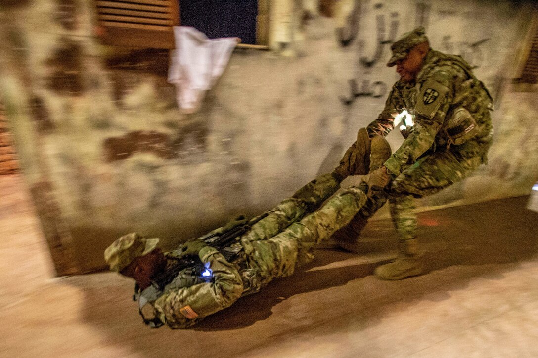 One soldier drags another by the legs on a cement-type floor near a wall with graffiti on it.