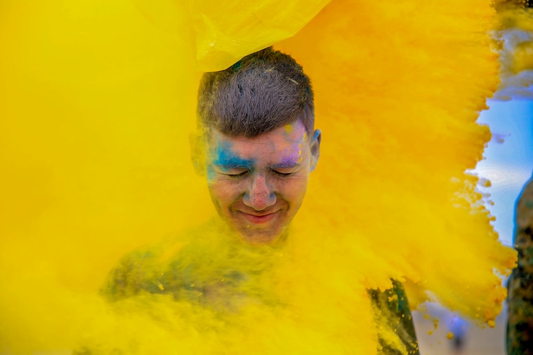 A Marine's grinning face, smeared with blue and purple powder, emerges from the center of a think cloud of yellow powder.