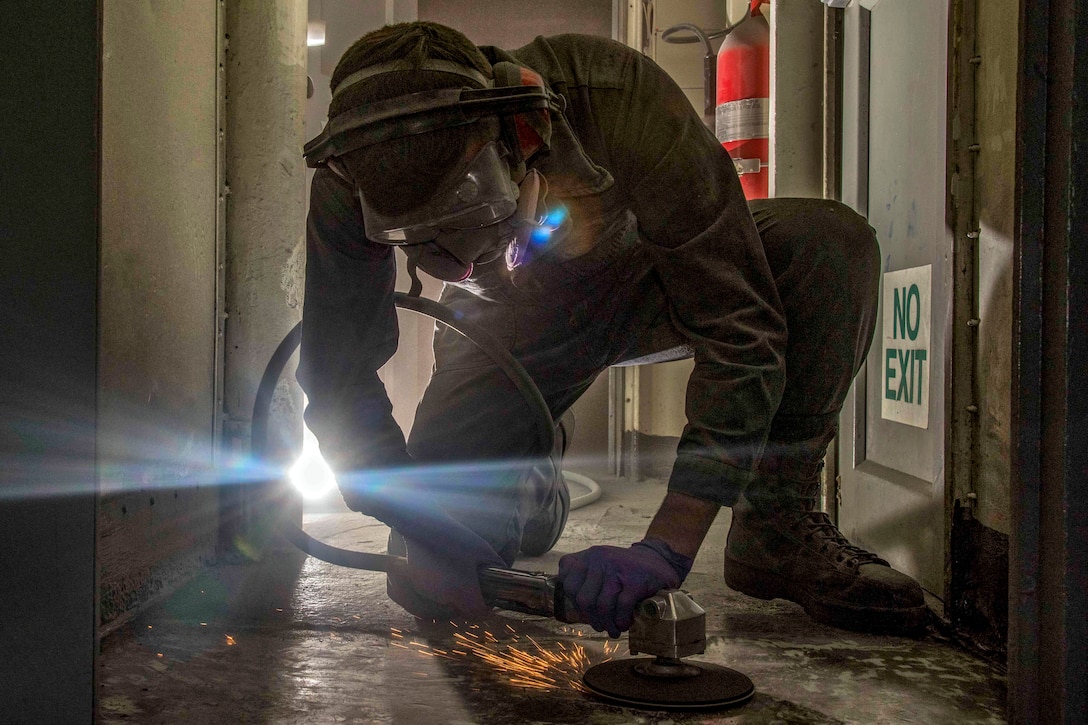 A sailor in protective head wear bends on one knee and uses a tool to grind a floor, creating sparks.