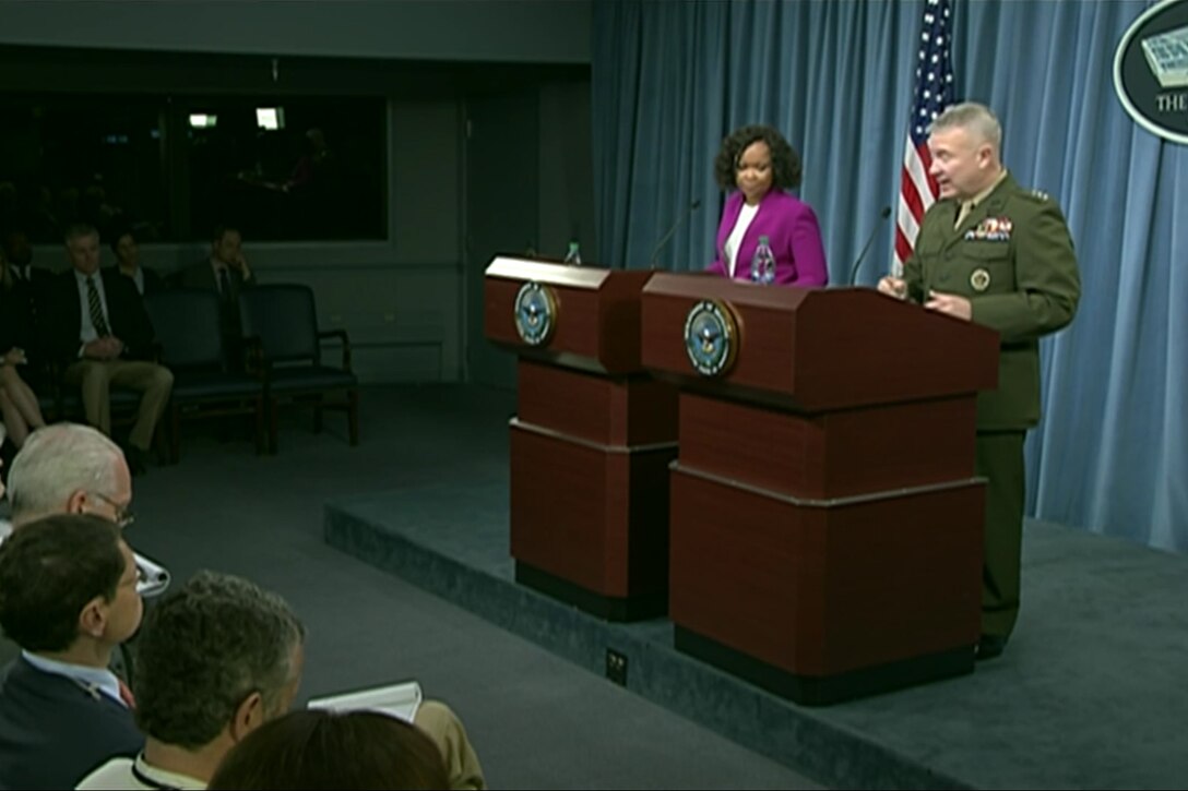 A civilian and a military official stand at lecterns and brief reporters.