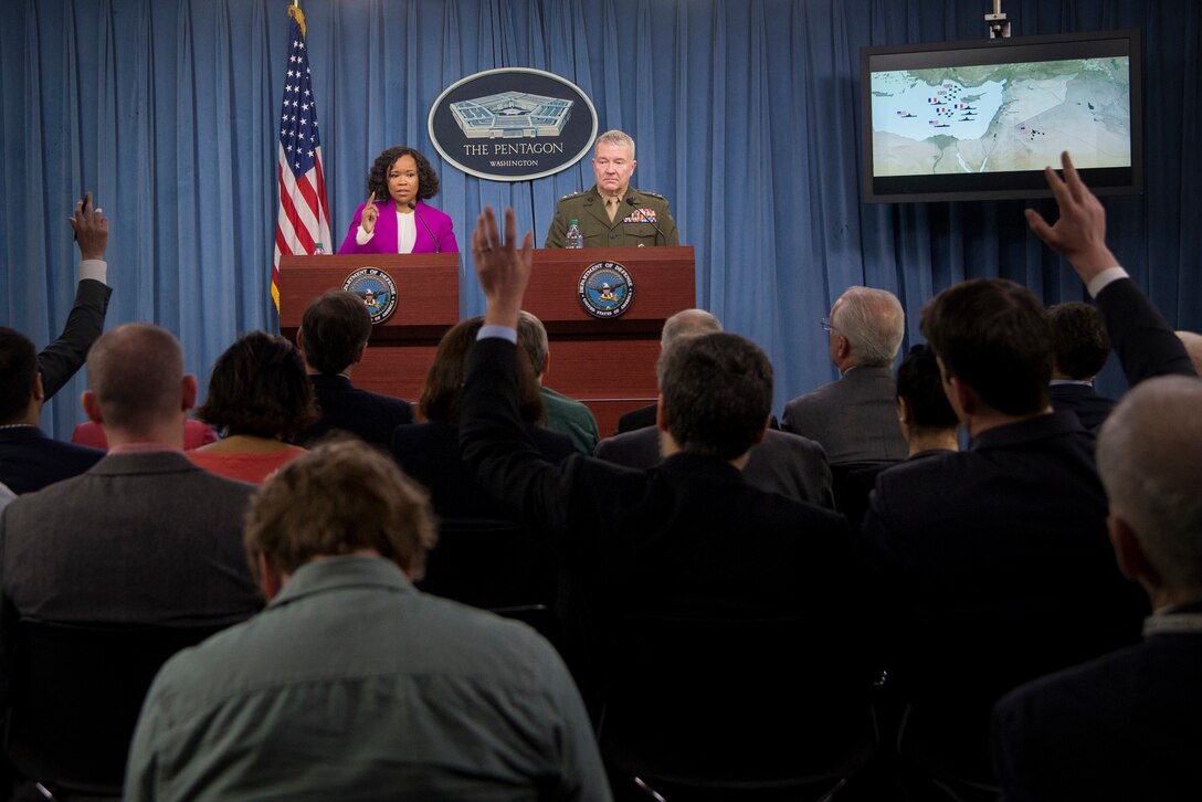 A civilian and a Marine stand behind lecterns and take questions from reporters.