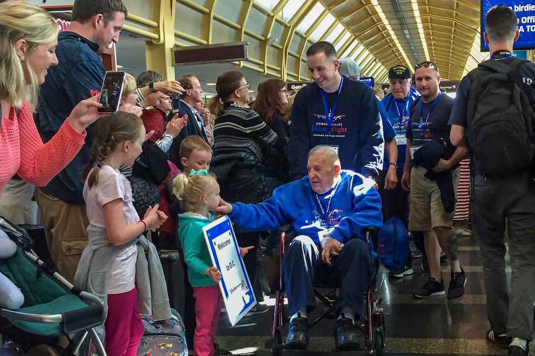 An elderly man in a wheelchair exchanges greetings with well-wishers gathered in an airport terminal.