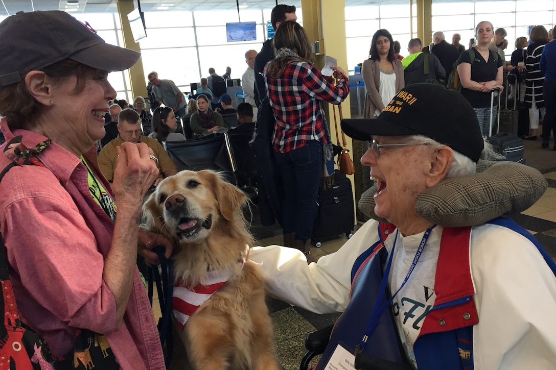 A woman and her dog greet a World War II veteran as he arrives at an airport.