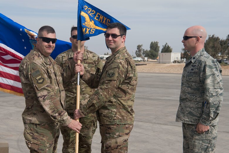 Maj. Robert Campbell, 332nd Expeditionary Maintenance Squadron incoming commander, accepts the unit guideon from Col. Shane Barrett, 332nd Expeditionary Maintenance Group commander, as Lt. Col. Scott May, 332nd EMXS outgoing commander, watches on April 8, 2018, at an undisclosed location in Southwest Asia.