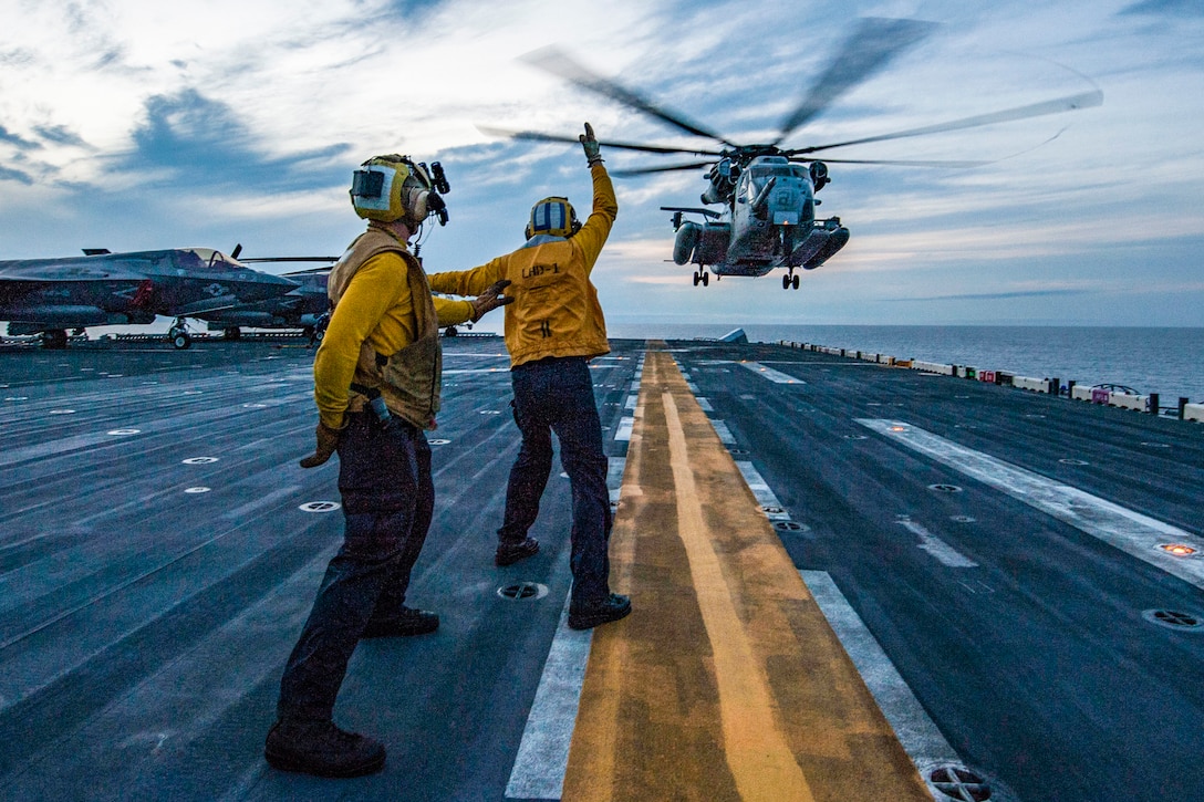 A helicopter lands on a ship with sailors using hand signals nearby.