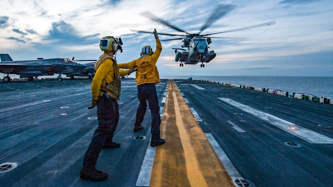A helicopter lands on a ship with sailors using hand signals nearby.
