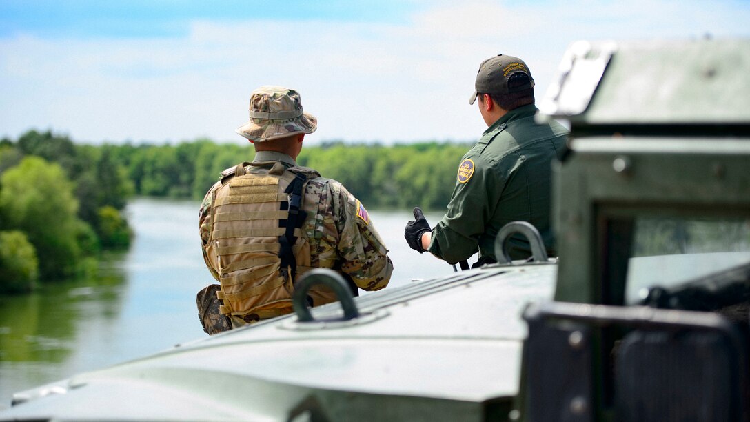 Two men in uniform stand next to a vehicle while looking toward a river.