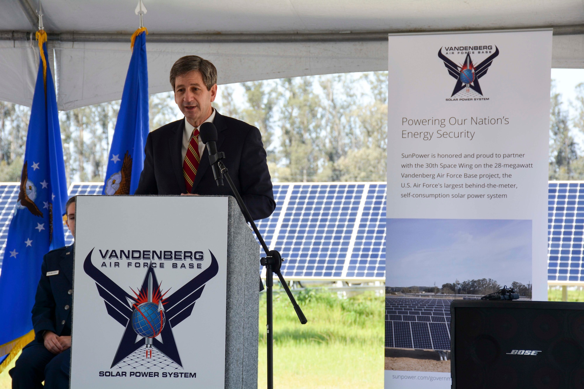 man speaks at podium on stage with rows of solar panels in background