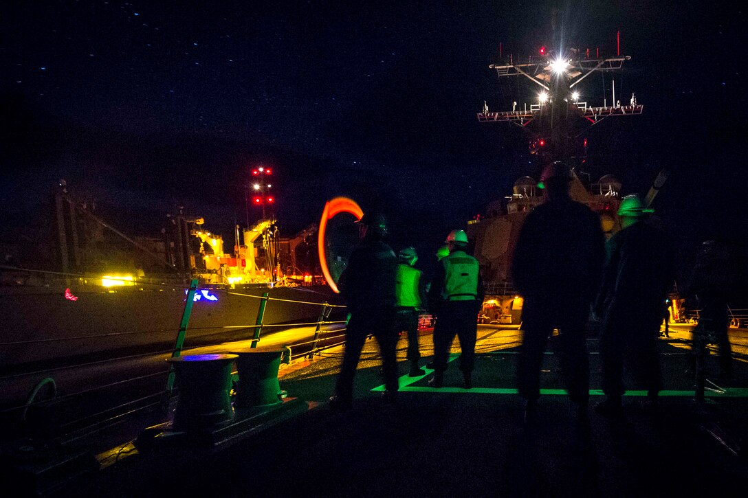 Sailors stand on the deck of a ship at night.