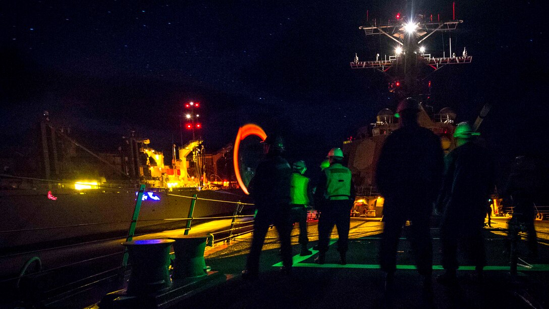 Sailors stand on the deck of a ship at night.