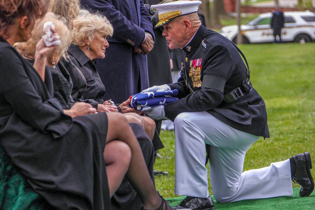 The commandant of the Marine Corps kneels to hand an American flag to a person.