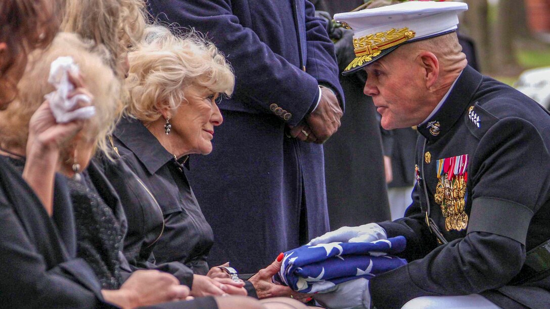 The commandant of the Marine Corps kneels to hand an American flag to a person.