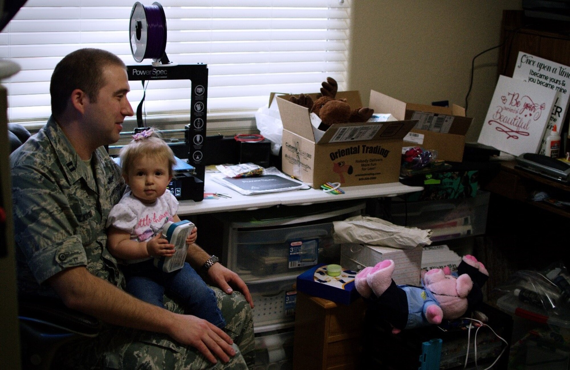 Capt. Jeremy Kulikowski, Air Force Life Cycle Management Center cyber mobile command and control systems officer, waits with his daughter, Kambri, for toy gastrostomy tubes to be 3-D printed so his wife, Kortni, can sew them onto stuffed animals, April 12, 2018. Their daughter, who was diagnosed with duplication syndrome when she was born in 2017, holds the pump to her old G-tube, which was removed from her stomach April 4, 2018. (U.S. Air Force photo by Audrey Jensen)