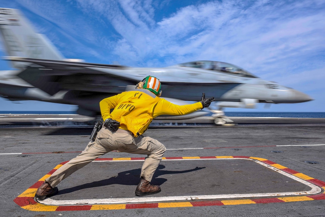 A sailor gestures while an aircraft moves in the background.