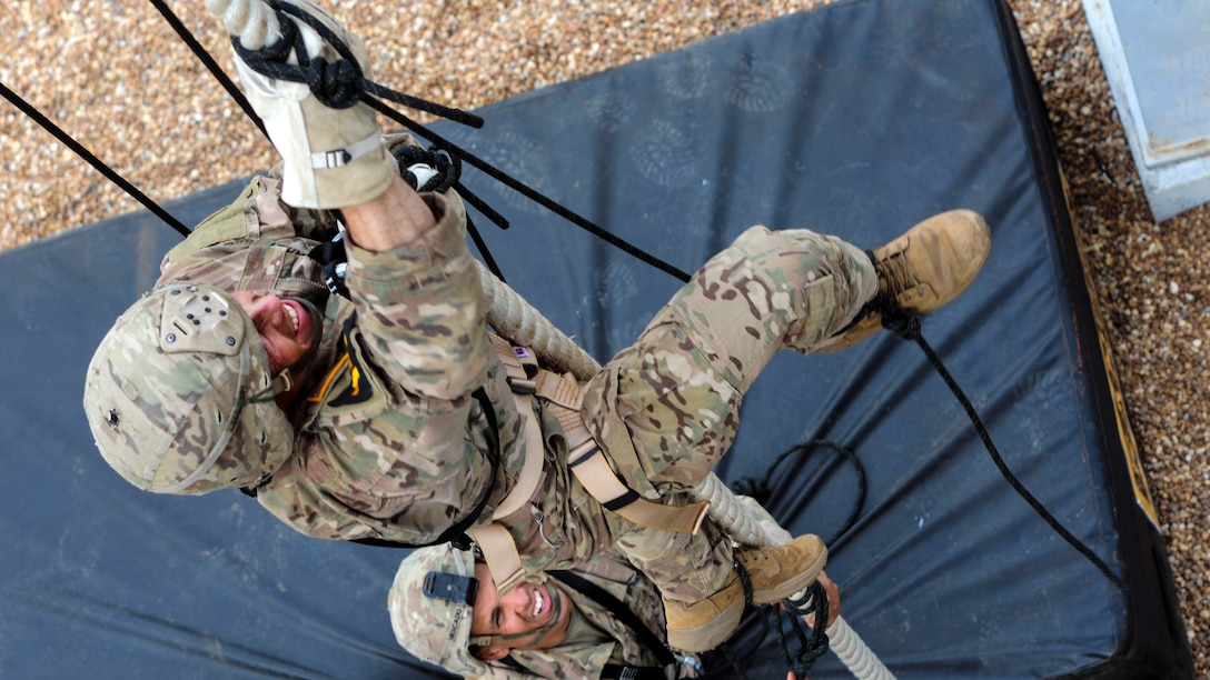 A soldier climbs a rope as another soldier watches from below.