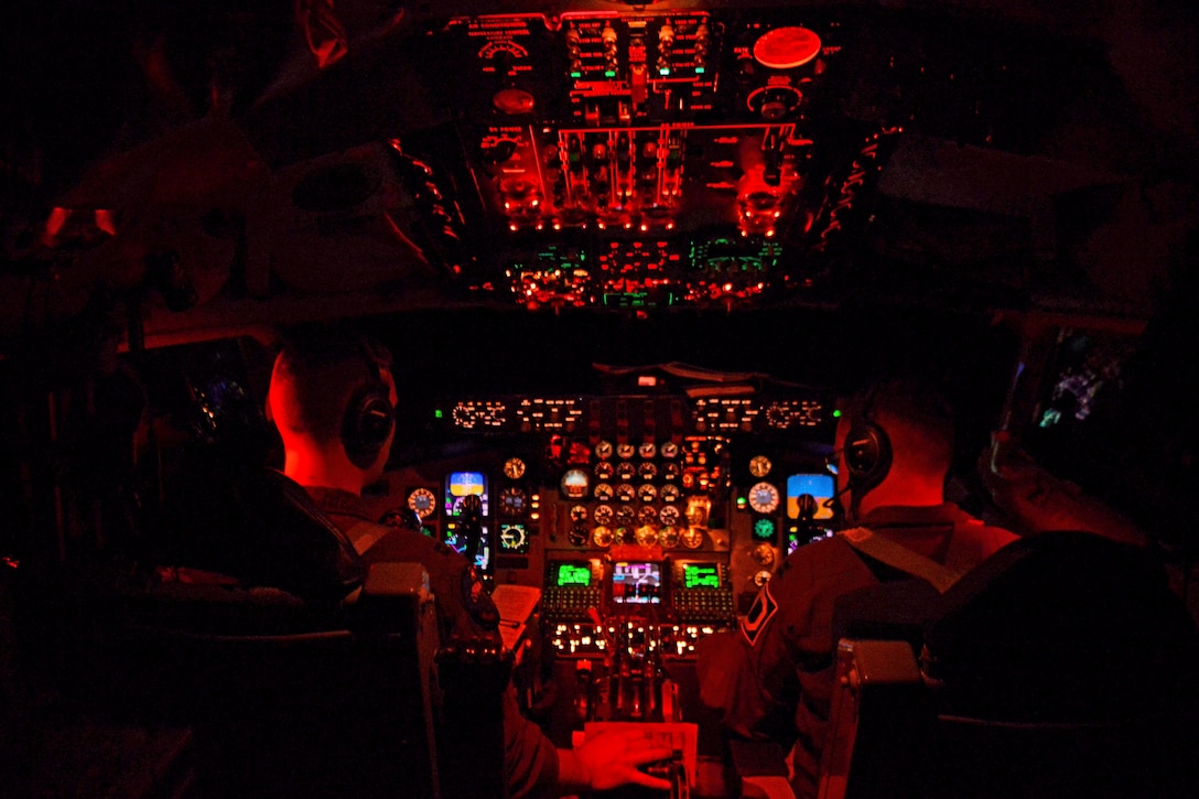 Red light casts a glow on the interior of a cockpit with two pilots at the controls.