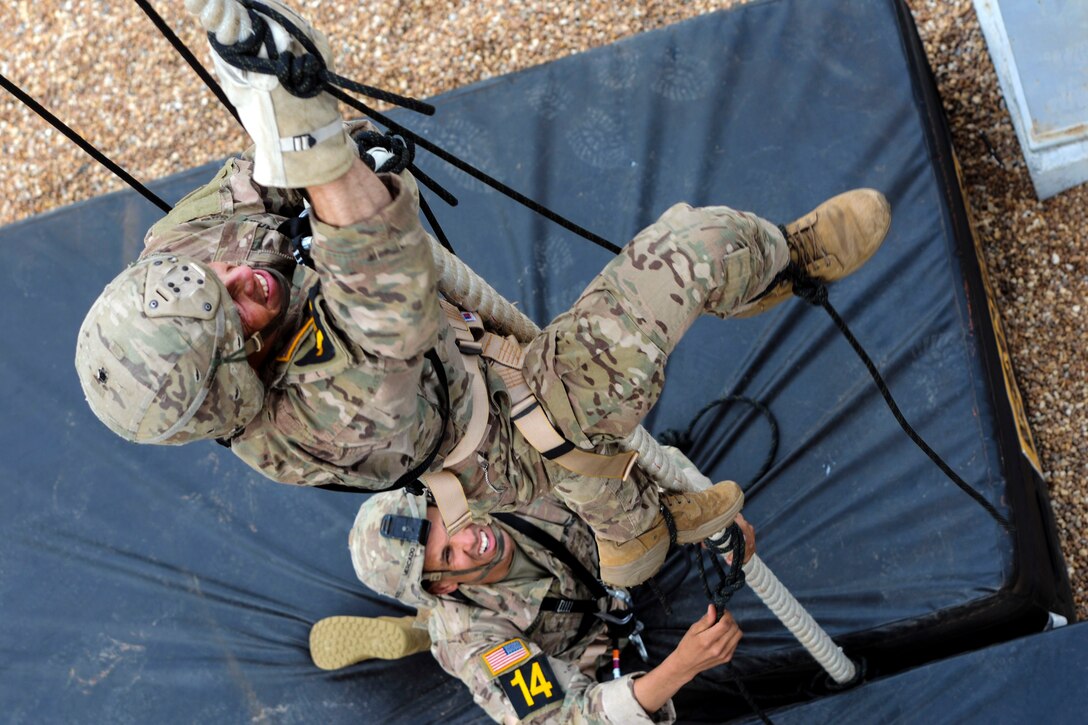 A soldier climbs a rope as another soldier watches from below.