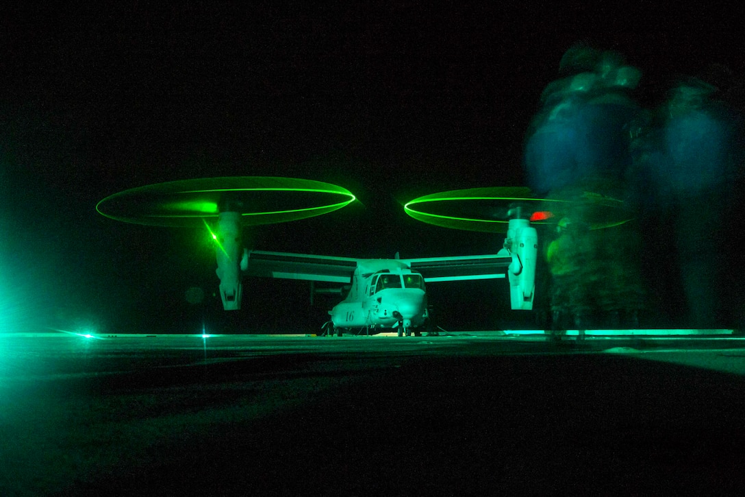 Service members stand on the deck of a ship as an aircraft lands.