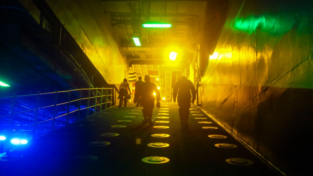 Two Marines walk up a ramp on a ship, with blue, yellow and green lights illuminating the path.