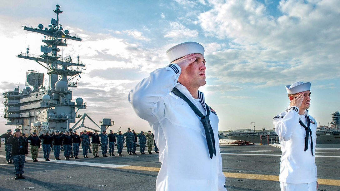 Two sailors salute on a ship's flight deck, as others gather behind them in the distance.