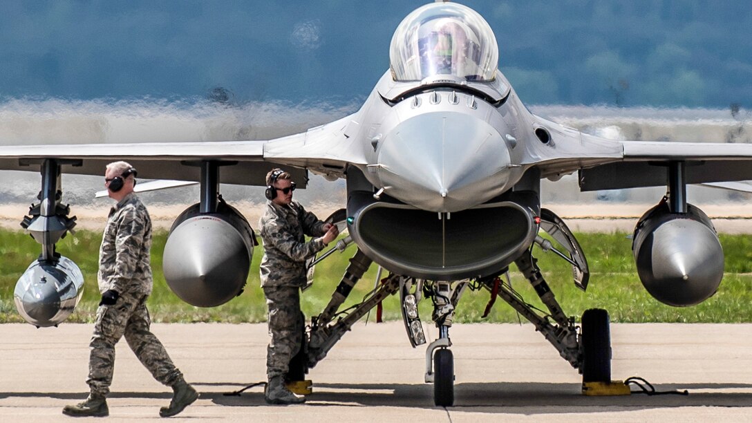 An airman works on a jet on a flightline as another airman walks nearby.